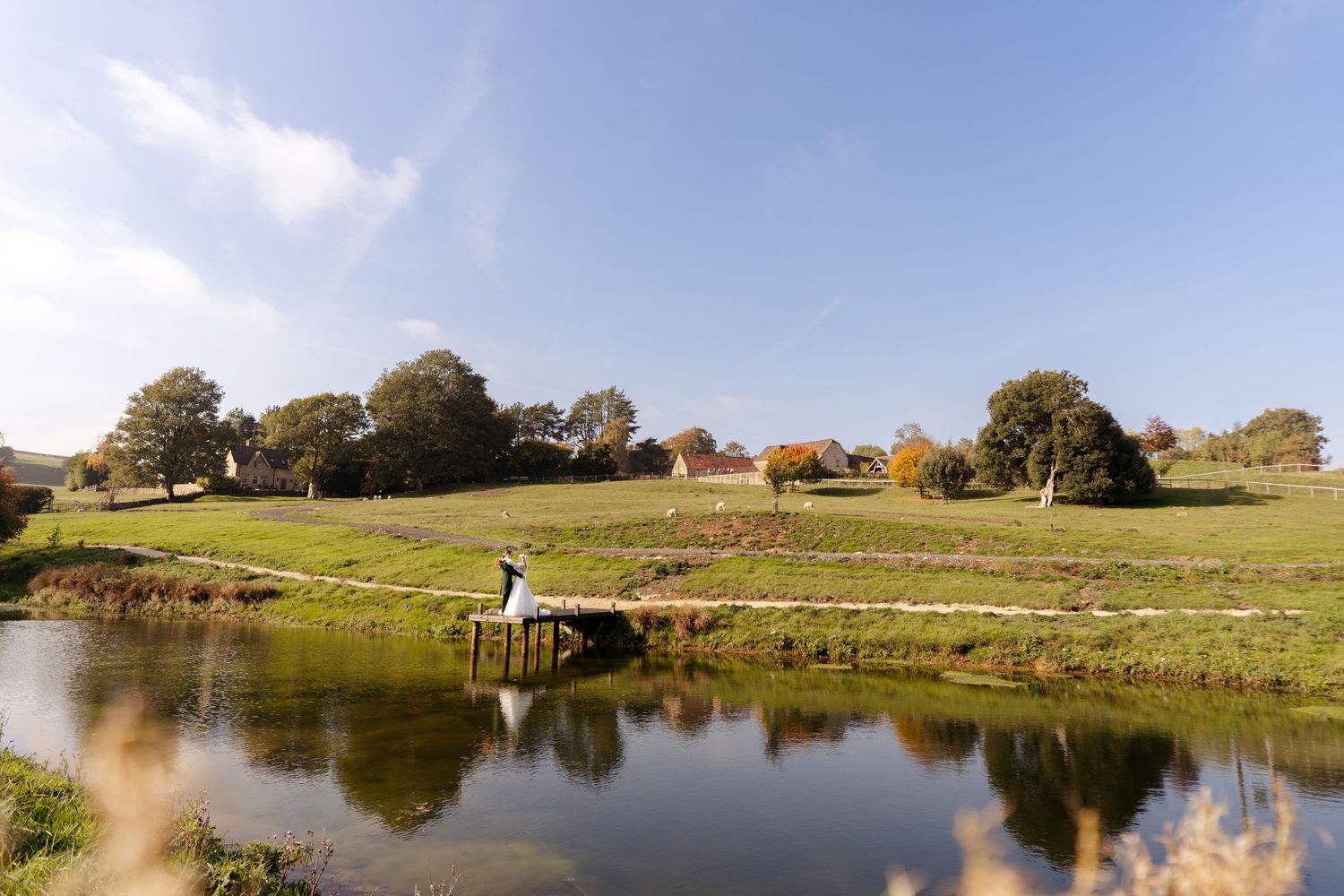 Bride sitting on a wooden dock by a serene pond surrounded by lush green hills and autumn trees on a sunny day.
