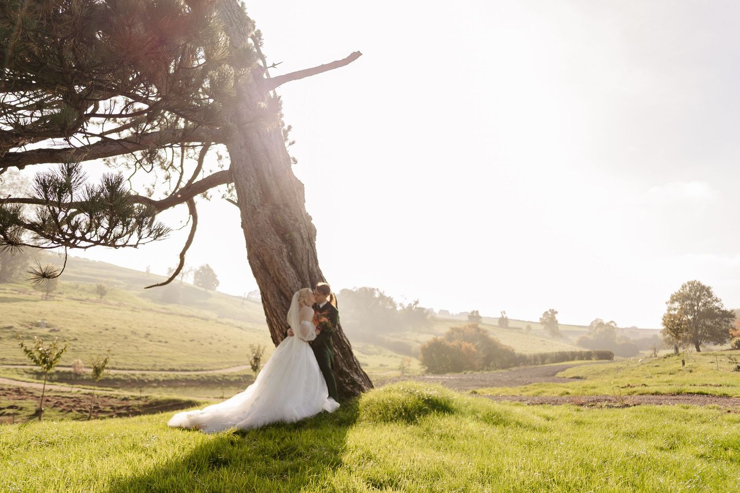 Bride and groom leaning against a tree in a sunlit green meadow, sharing a romantic moment in wedding attire.