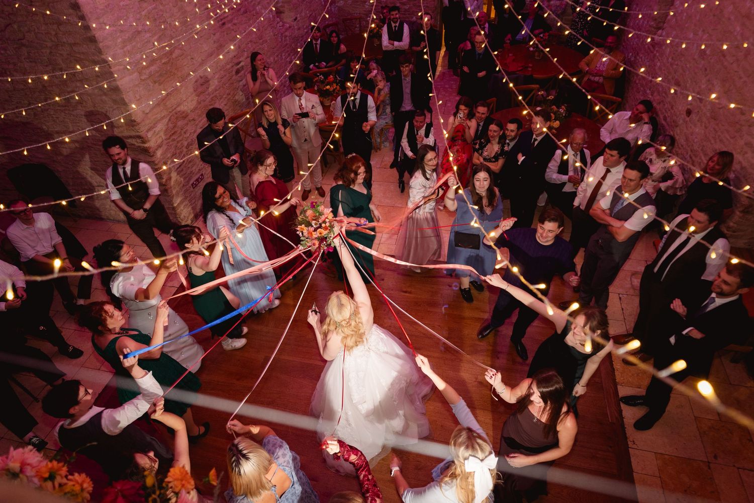 Aerial view of wedding reception dance floor with guests holding colorful ribbons around the bride in a festive tent venue.