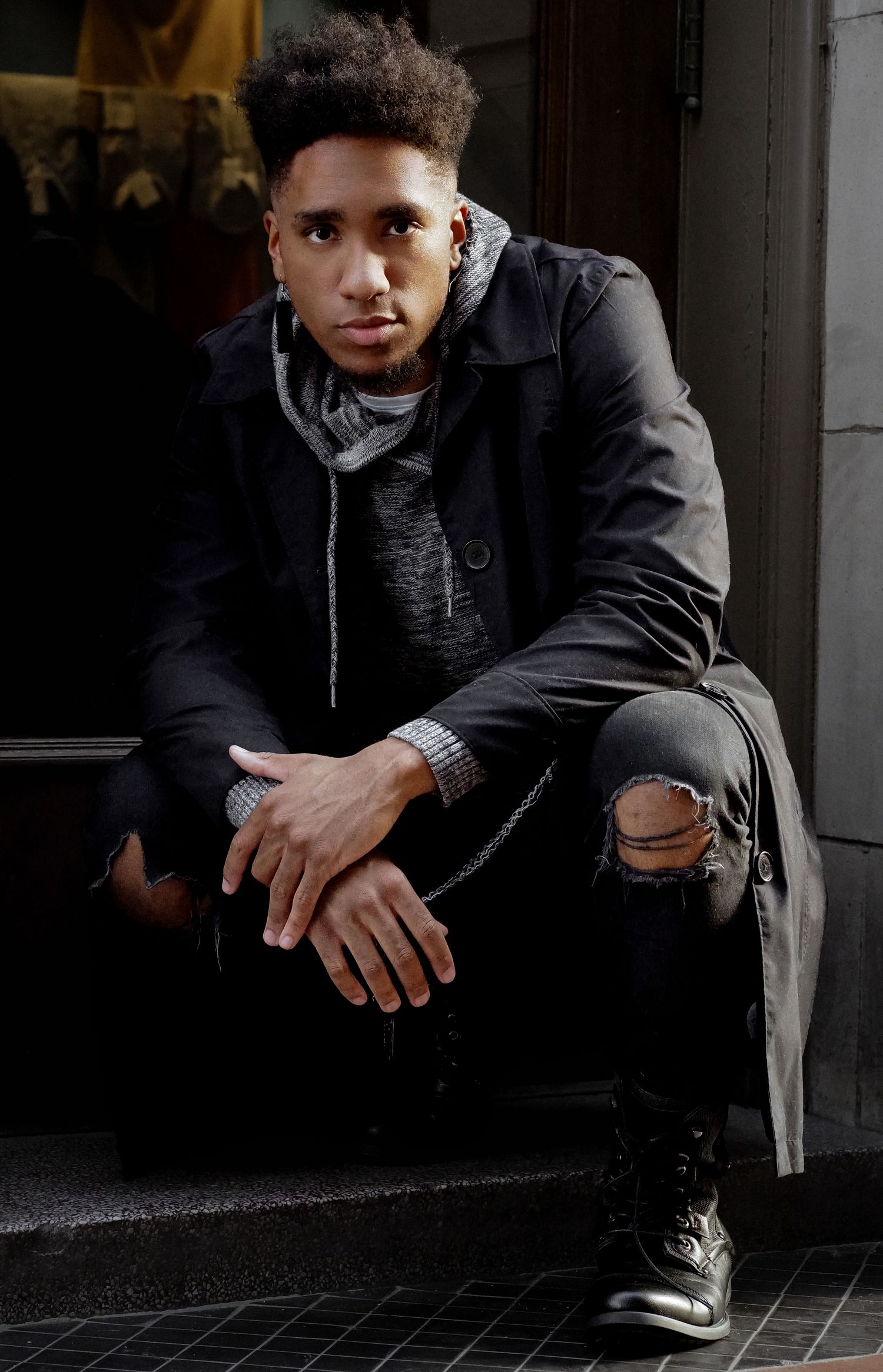 Young Black man in ripped jeans and black coat sits on steps, posing confidently for a urban fashion portrait.