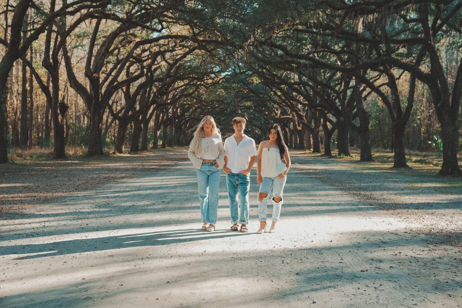 Family of four walking together on a tree-lined path in matching white tops and jeans on a sunny day.