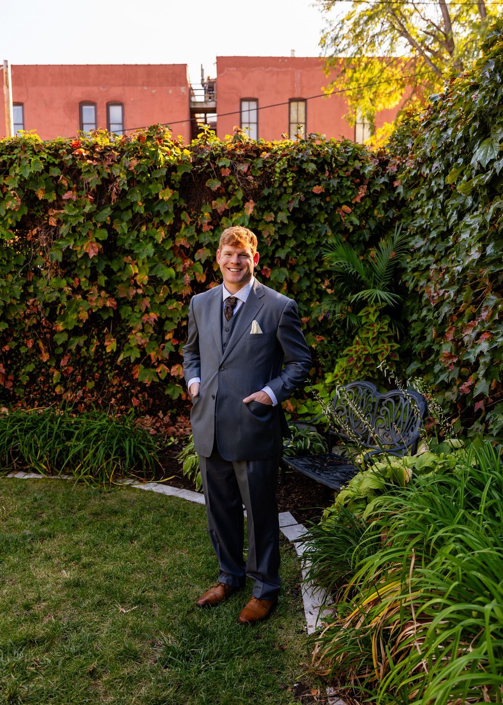 Groom in gray suit standing in lush garden with ivy-covered wall at outdoor wedding venue.
