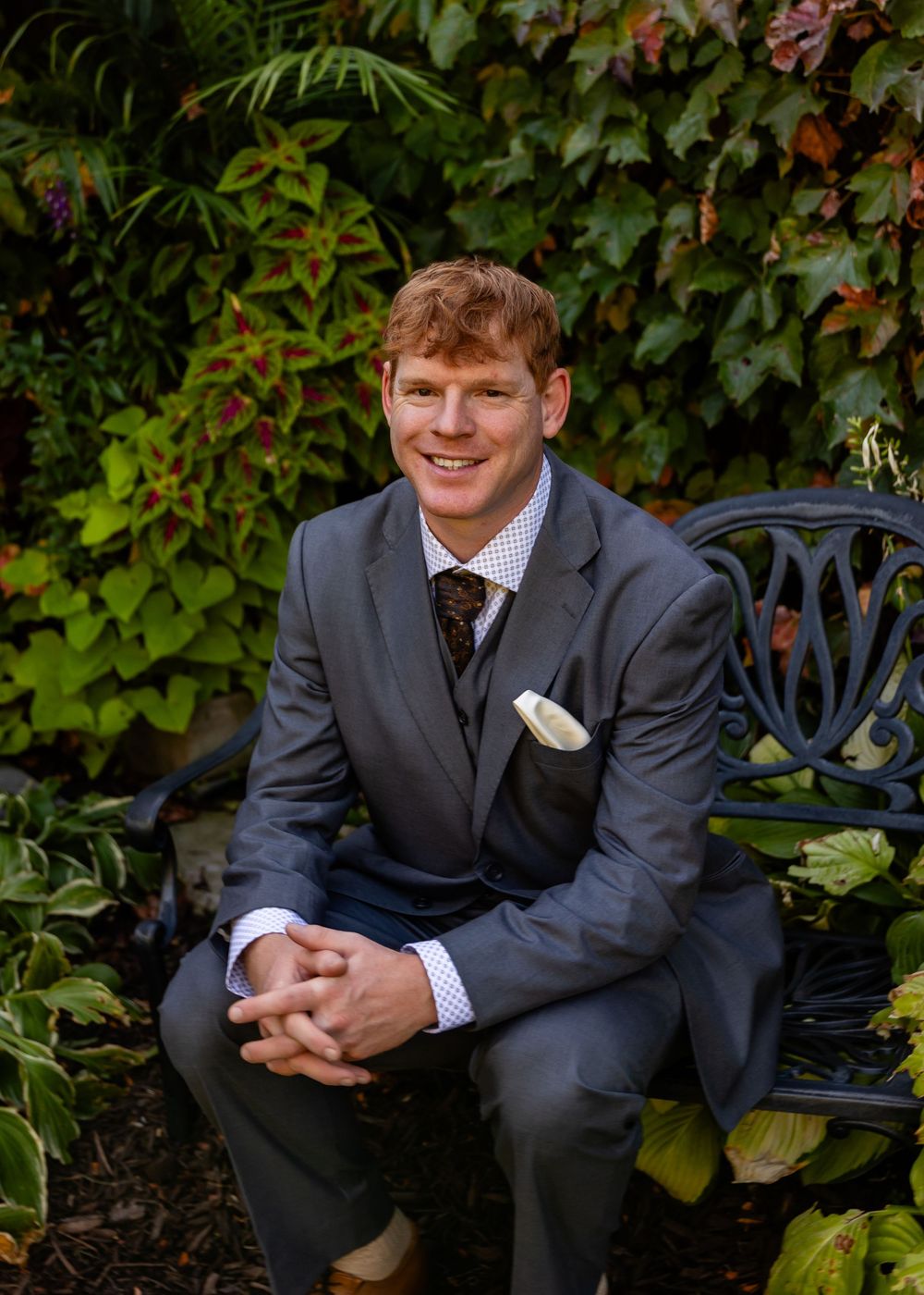 Dapper redhead groom in gray three-piece suit sitting on ornate bench with colorful autumn garden backdrop.