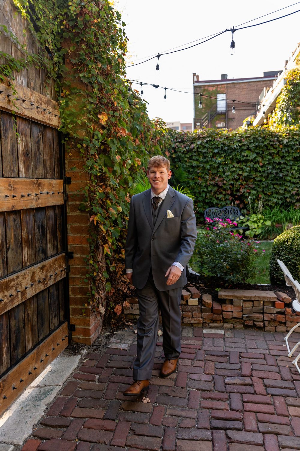 Groom in gray suit walks brick pathway through lush ivy-covered garden walls at outdoor wedding venue.