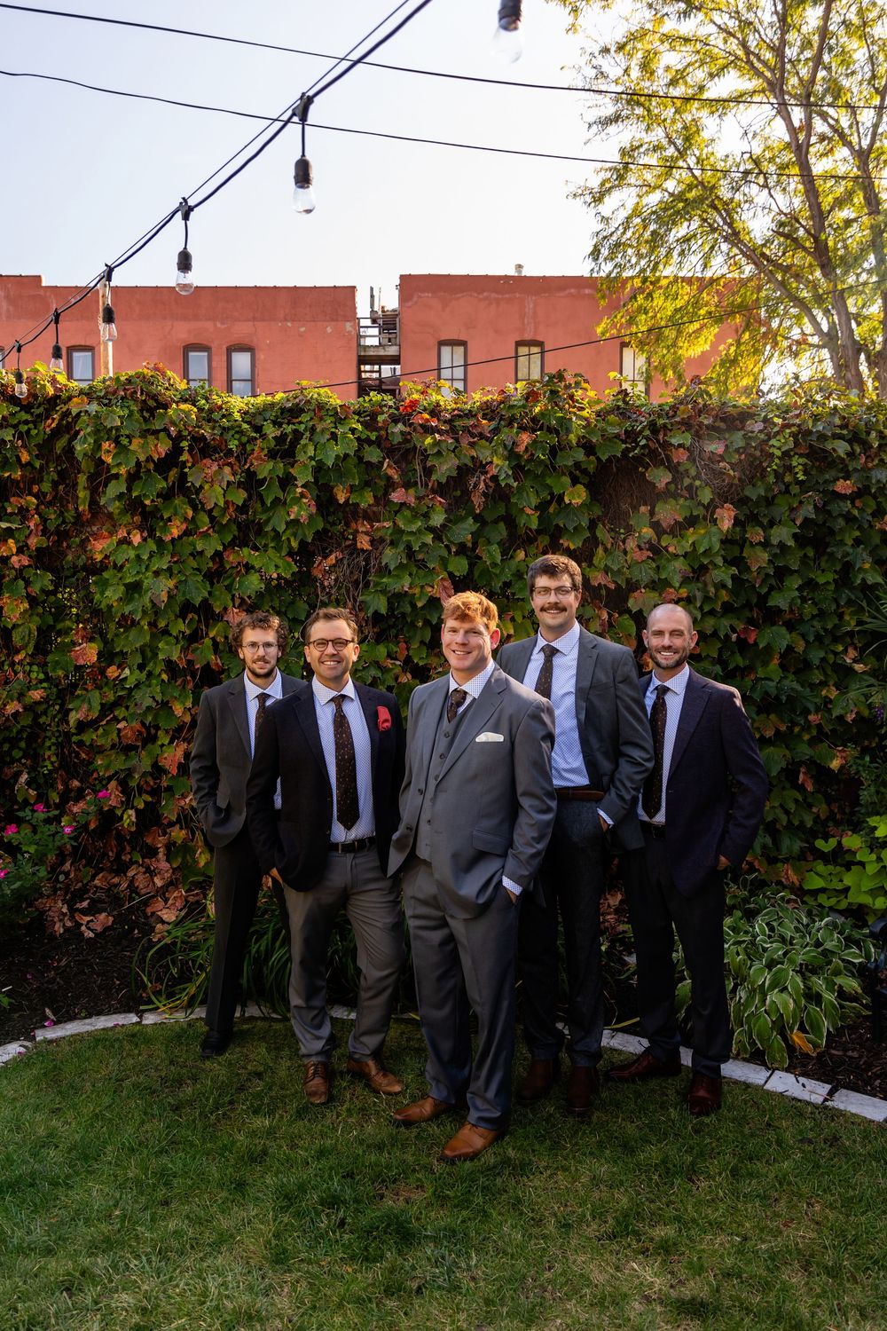 Five groomsmen in suits pose together in a lush ivy-covered garden at golden hour.