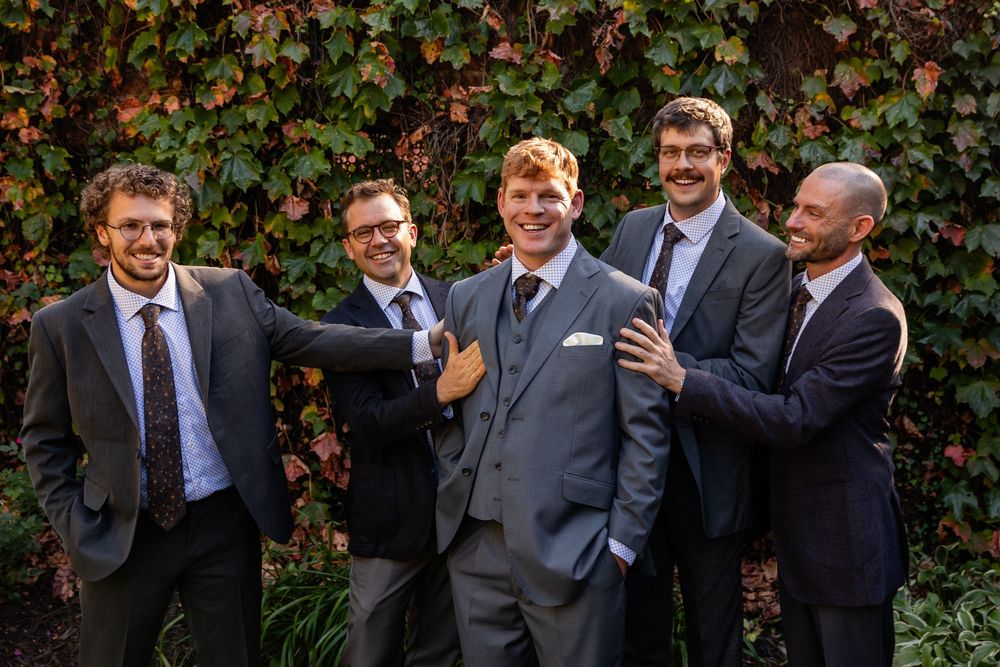 Five groomsmen in suits laughing together outdoors against a lush ivy-covered wall at a wedding.