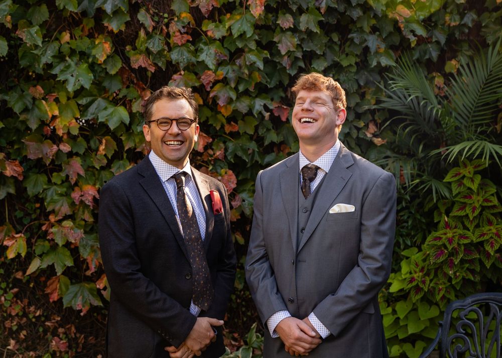 Two men in suits smiling outdoors against a lush ivy-covered wall at a wedding celebration.