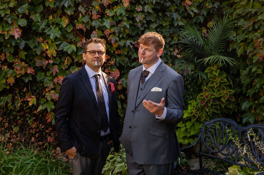 Two men in suits pose outdoors against a lush ivy-covered wall at a formal event.