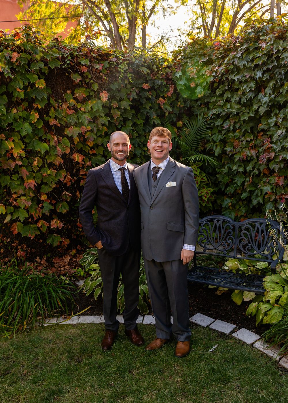 Two grooms in suits pose together in a lush garden with ivy-covered walls and an ornate bench.