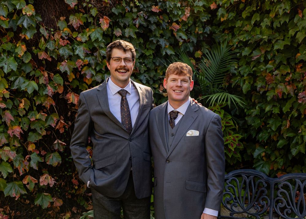 Two groomsmen in gray suits smile together in front of colorful autumn ivy wall at wedding.