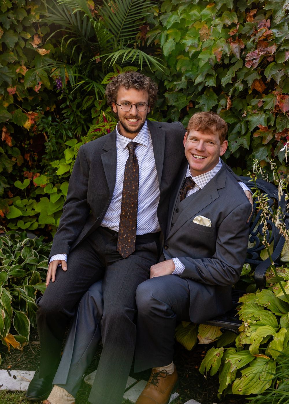 Two men in suits smiling together seated among green foliage at an outdoor wedding.
