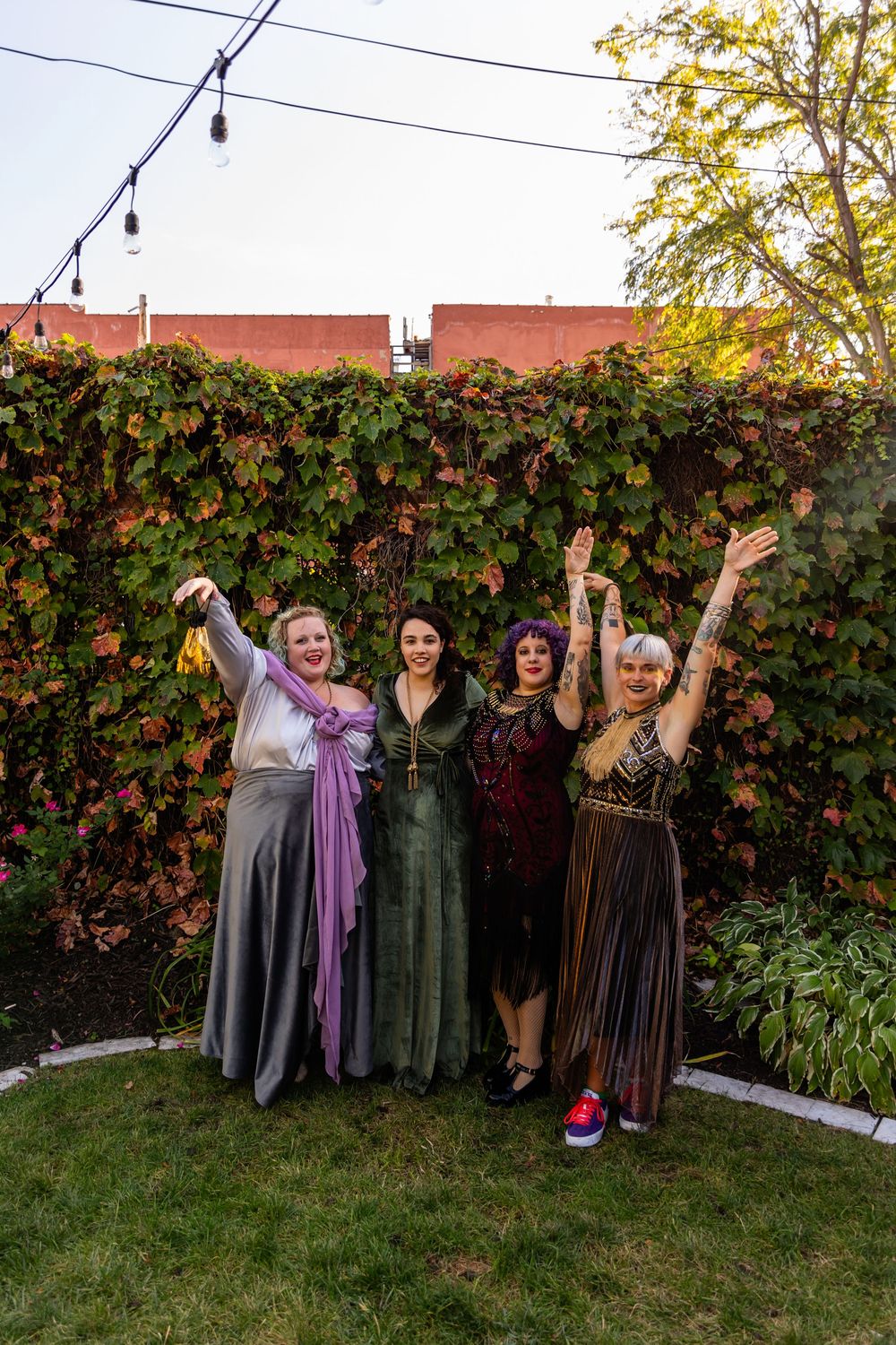 Four women in elegant colorful dresses celebrating with raised arms at outdoor wedding reception.