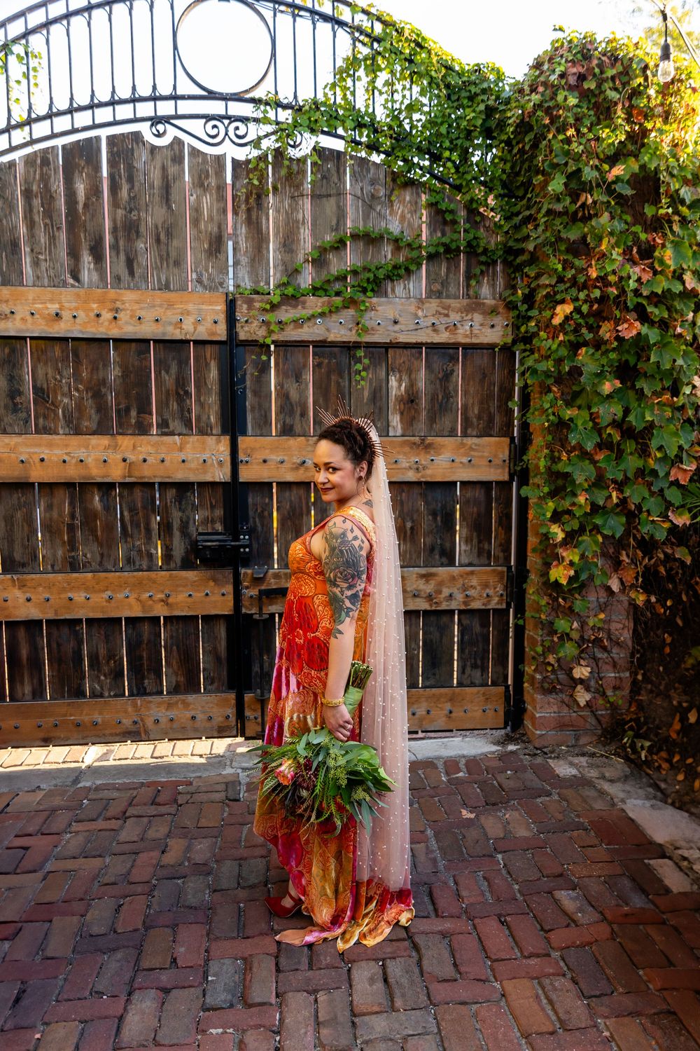 Bride in colorful orange wedding gown with veil poses by rustic wooden gate covered in autumn ivy.