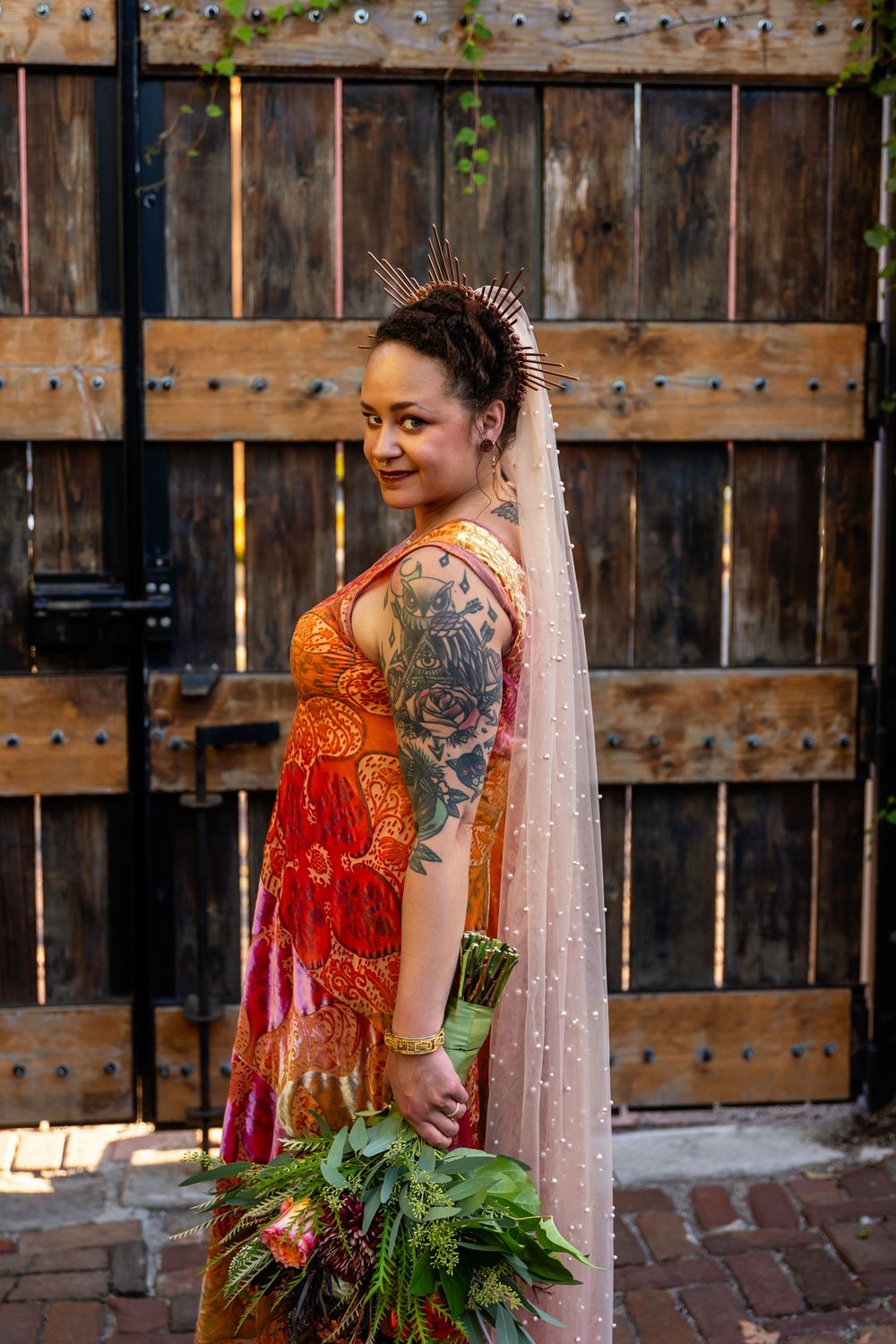 Bride in vibrant red-orange gown with veil and floral crown poses by rustic wooden doors.