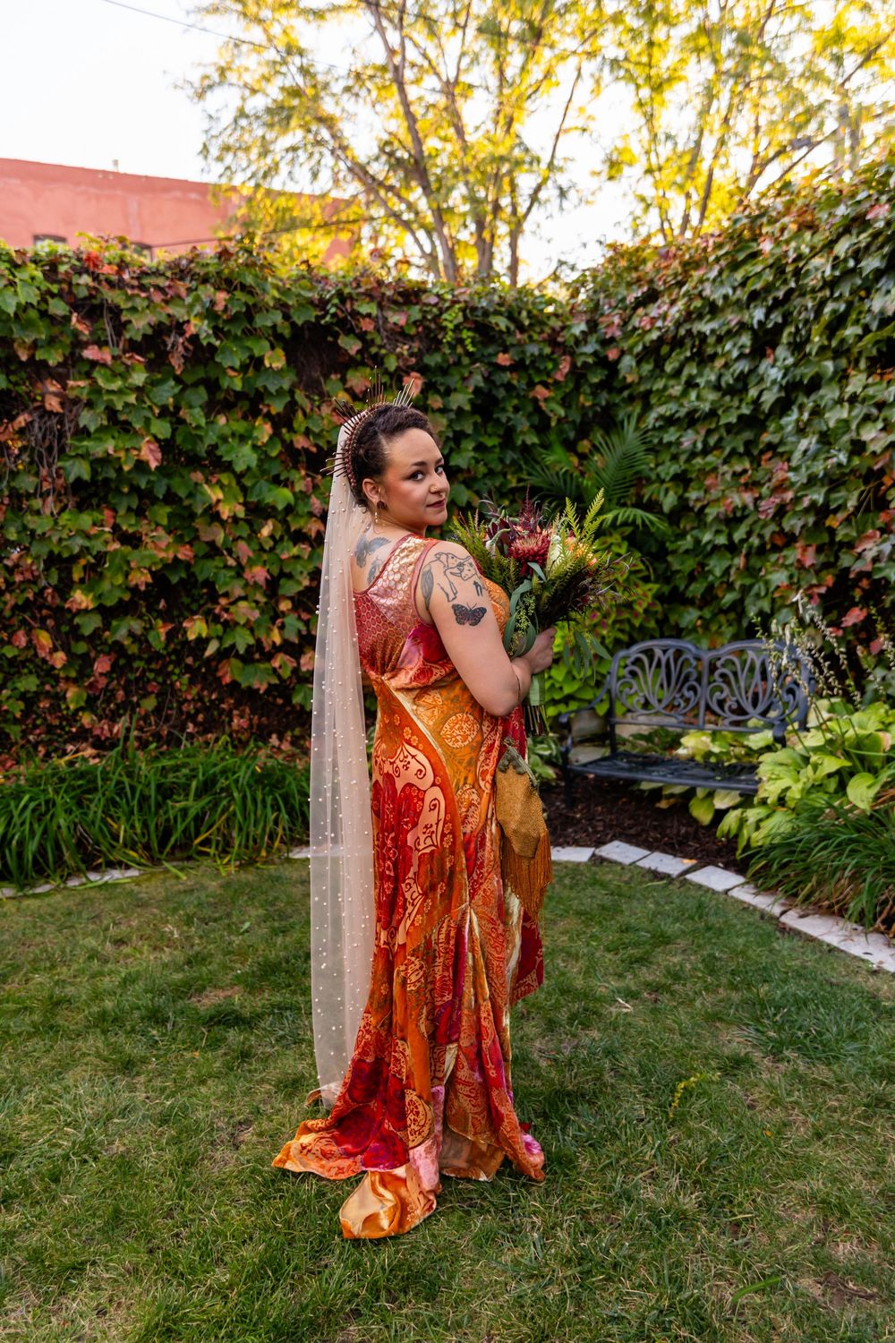 A bride in a vibrant orange and red sari holds a bouquet while standing in a lush garden with ivy-covered walls.