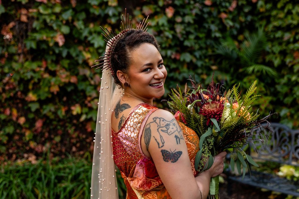 Tattooed bride in orange dress smiles over shoulder wearing crown headpiece with autumn bouquet.