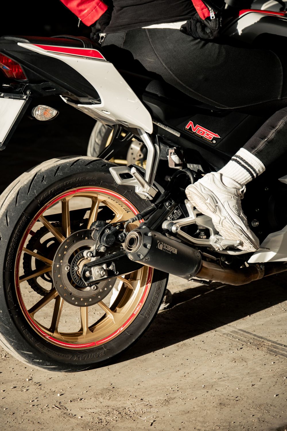 Close-up of a sport motorcycle rear wheel with gold rims and exhaust pipe, rider wearing white sneakers visible.