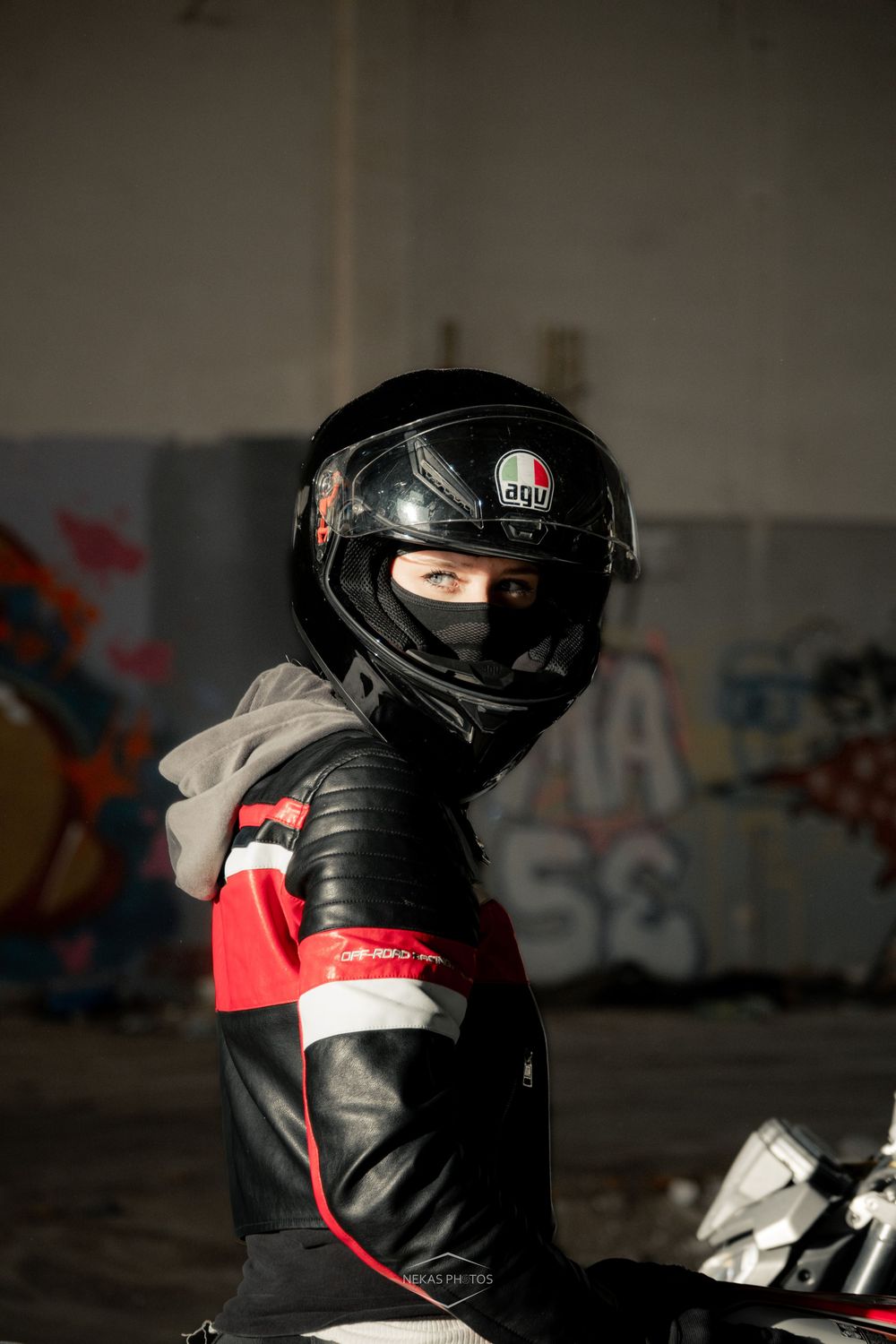 Motorcyclist wearing a black AGV helmet and leather racing jacket sitting on a bike in front of graffiti wall.