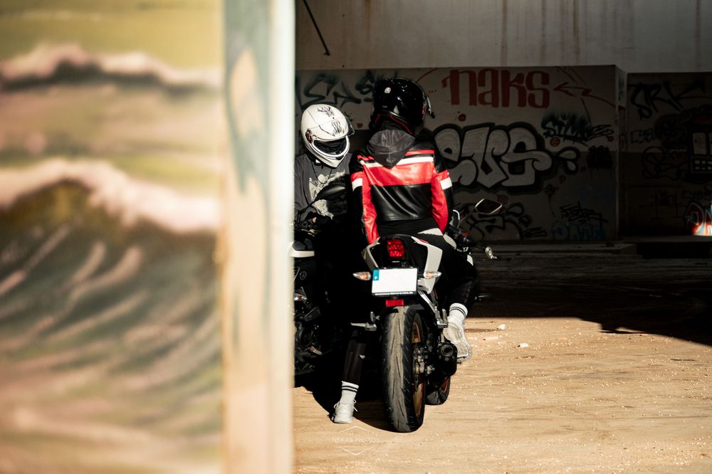 Motorcyclist in red jacket and helmet parked near graffiti-covered wall in urban setting.