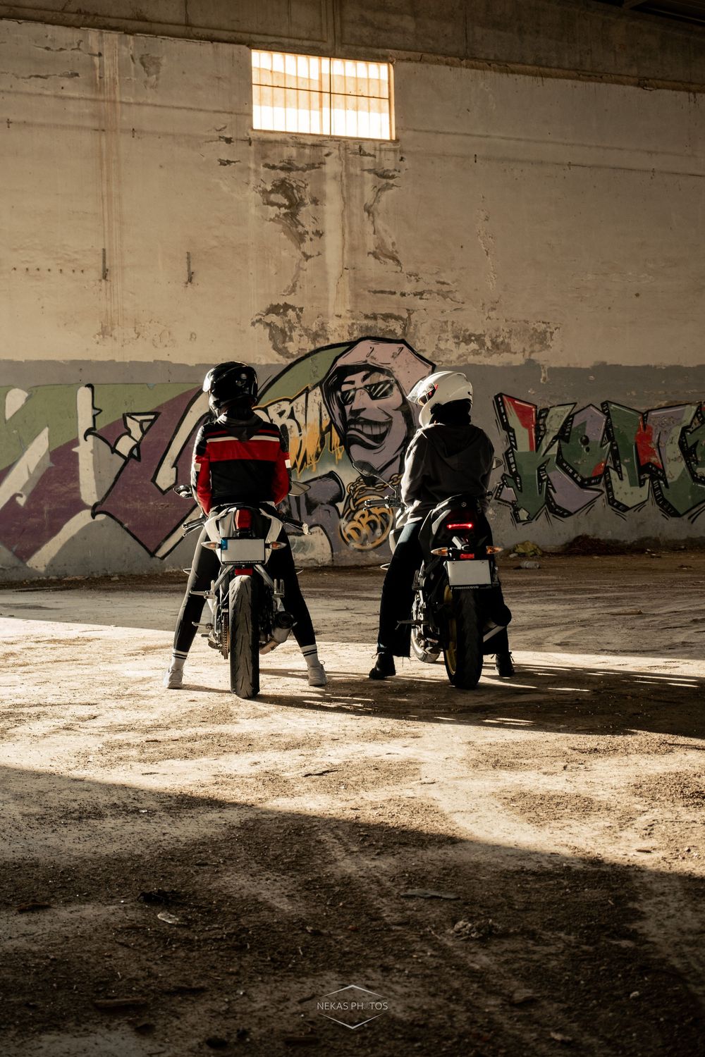 Two motorcyclists in helmets sit on bikes inside an abandoned building with colorful graffiti wall art in the background.