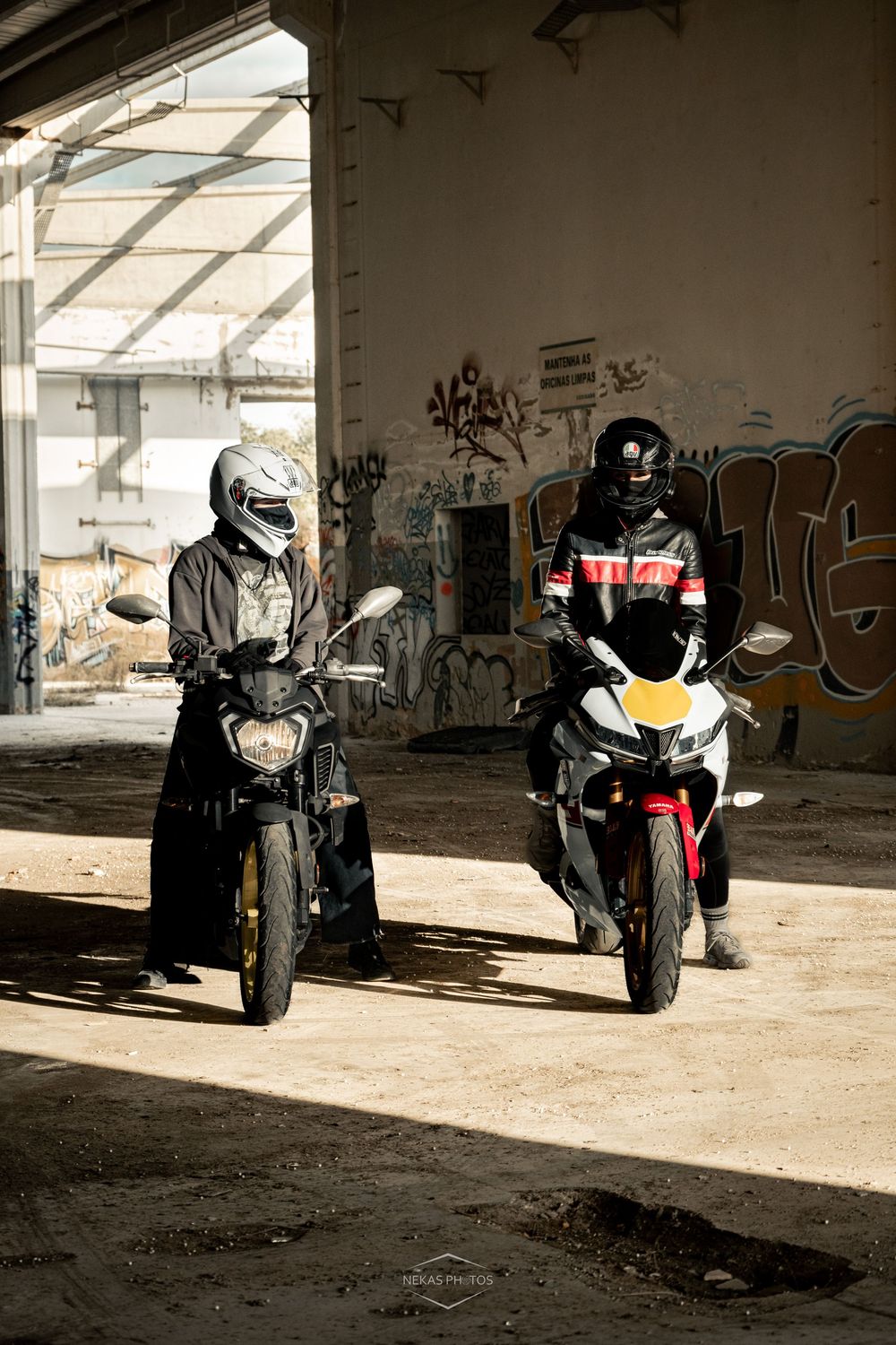 Two motorcyclists in helmets riding bikes inside an abandoned graffiti-covered building with sunlight streaming in.