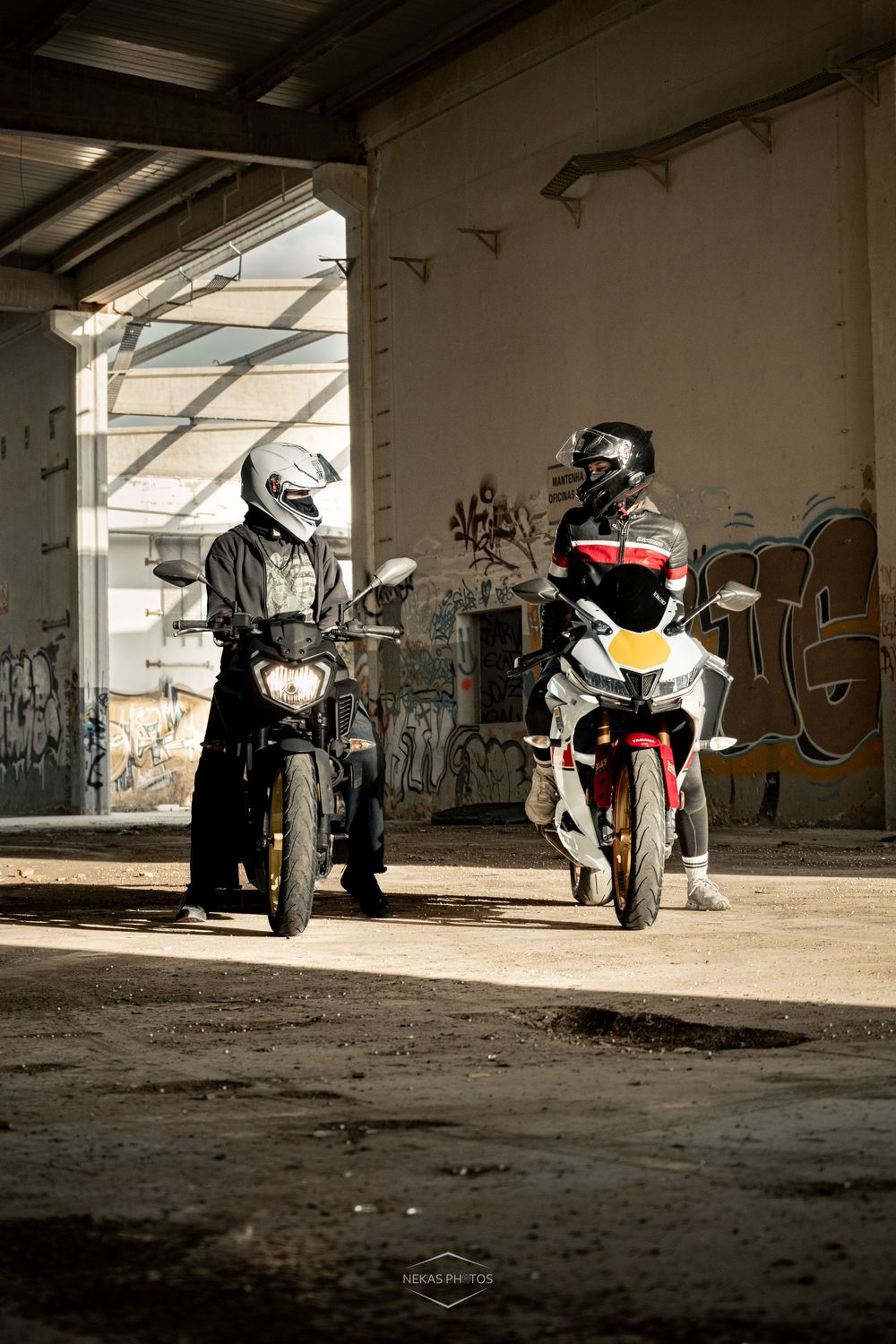 Two motorcyclists in helmets and riding gear pose on their bikes inside an abandoned building with graffiti walls.