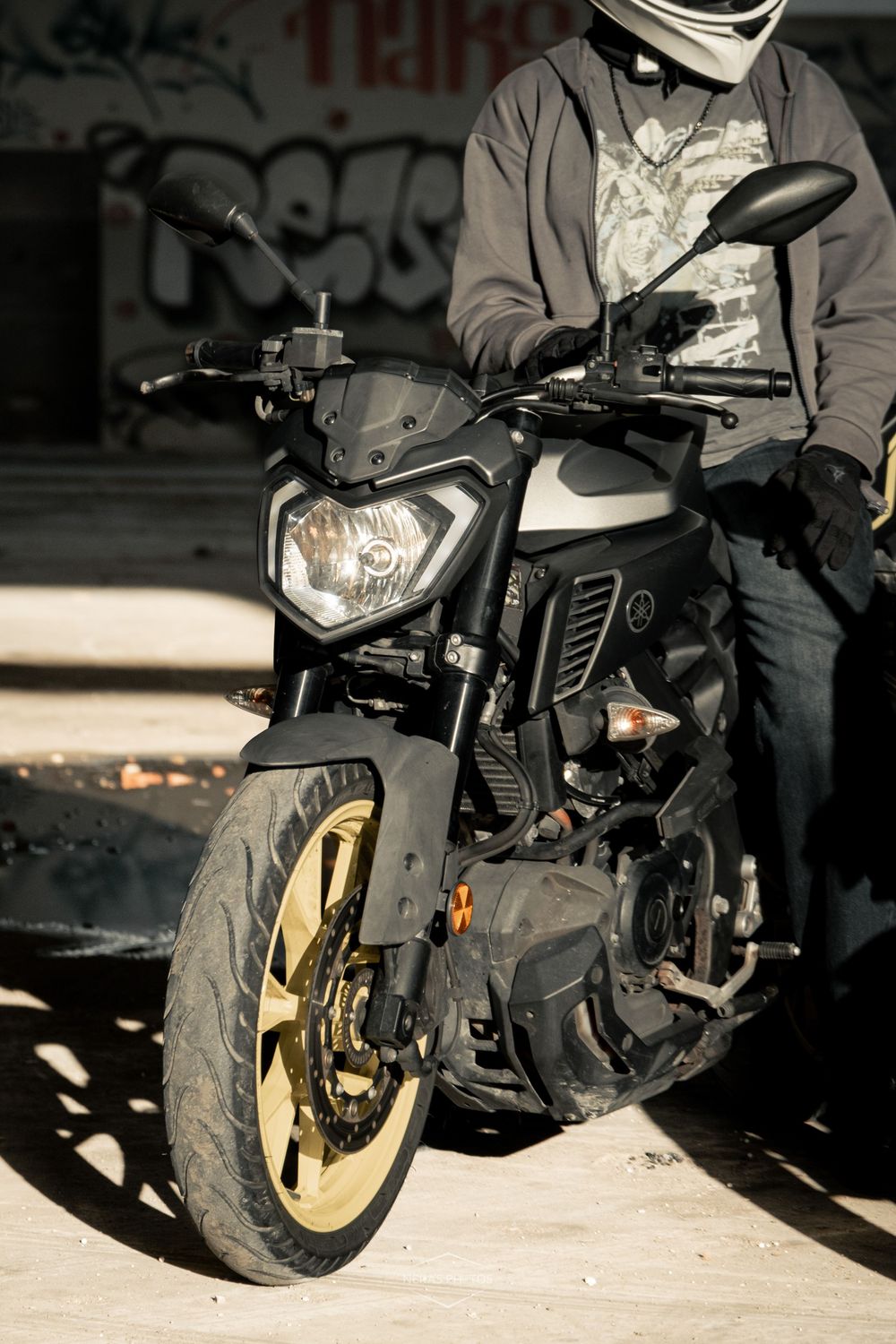 A motorcyclist in a helmet stands beside a black and gold Yamaha MT motorcycle near a graffiti wall.