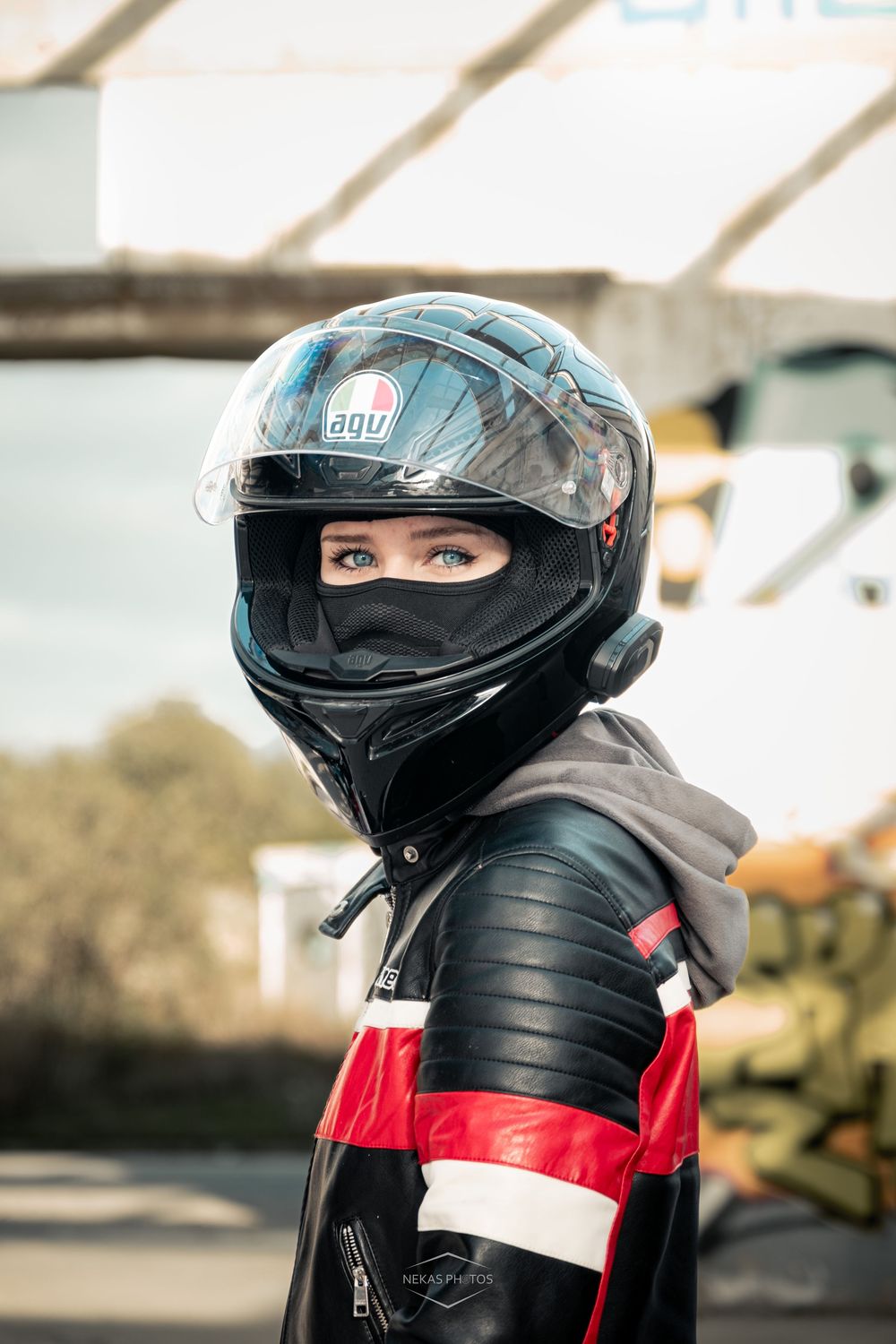 A female motorcyclist wearing an AGV helmet and black red leather racing jacket poses outdoors at a track.