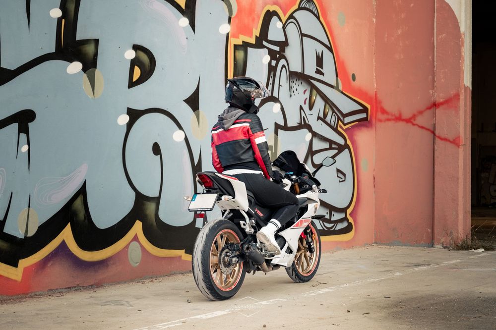 Motorcyclist in red jacket and helmet parked near colorful graffiti wall on a white sport bike.