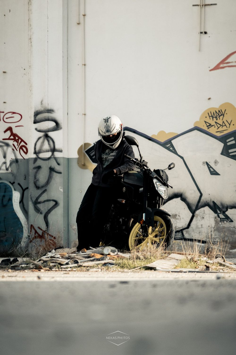 Motorcyclist in black gear and white helmet posed on a bike against a graffiti-covered urban wall.