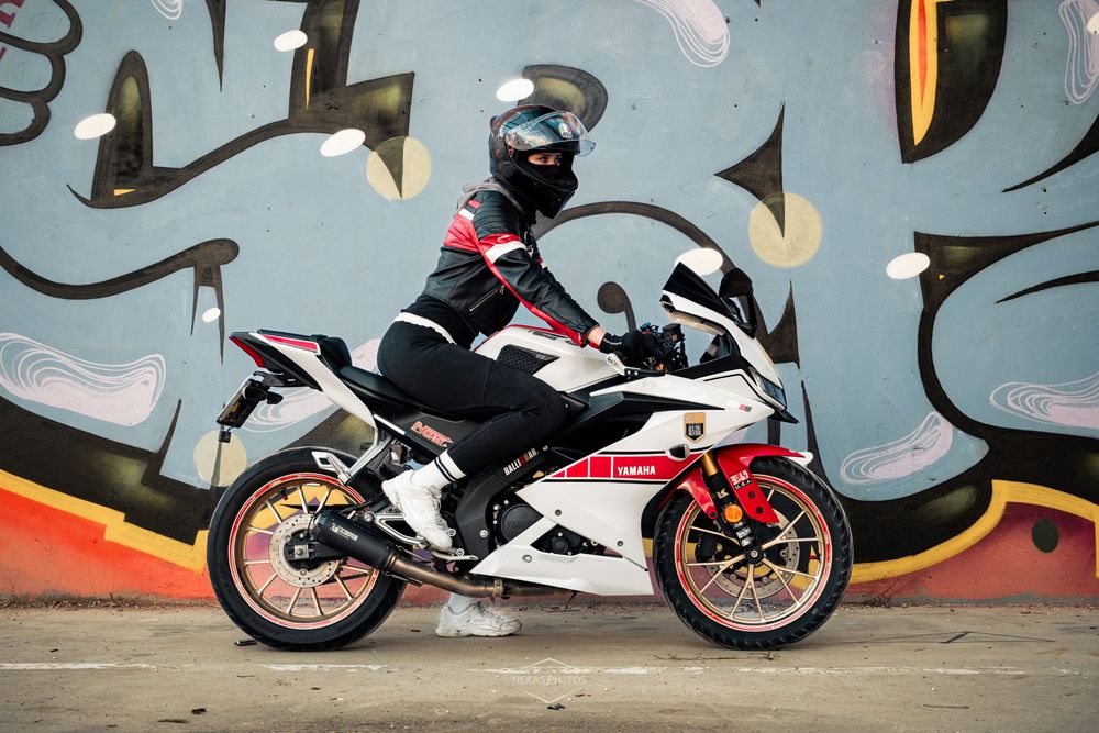 A motorcyclist in black gear rides a white and red sport bike in front of a colorful graffiti wall.