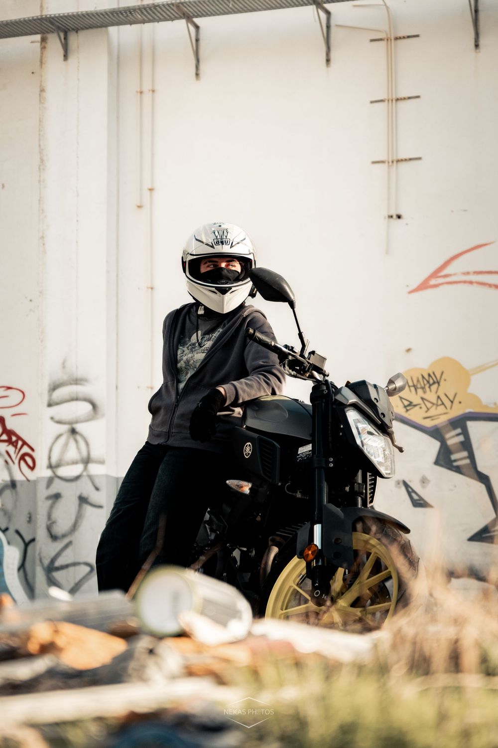 A motorcyclist wearing a white helmet sits on a dark motorcycle in front of a graffiti-covered wall.