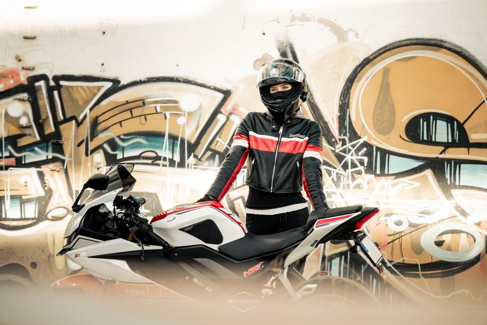 Motorcyclist in full racing gear and helmet poses on a white sport bike in front of colorful graffiti wall.
