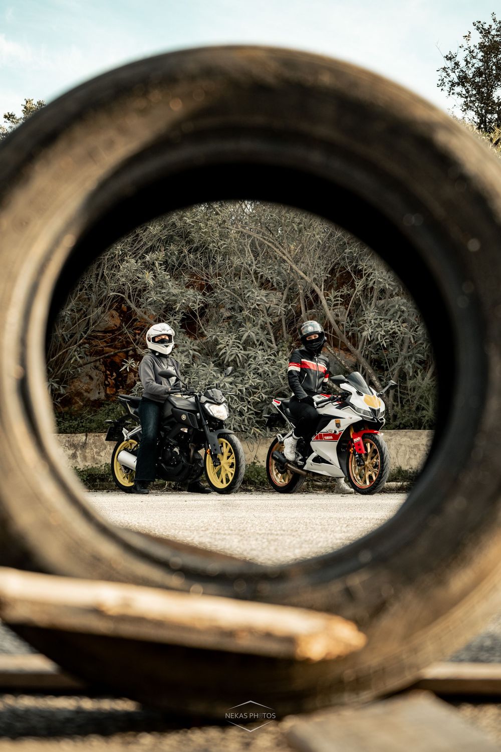 Two motorcyclists with helmets posed on sport bikes, viewed through a tire frame, with trees in the background.