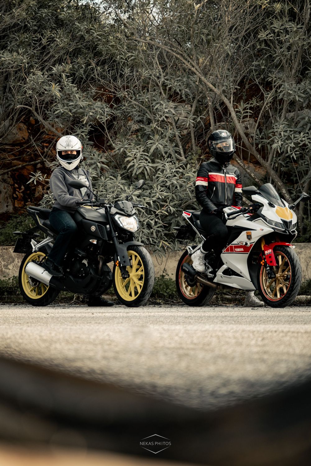 Two motorcyclists in helmets pose on sport bikes with gold wheels on a road surrounded by lush trees.