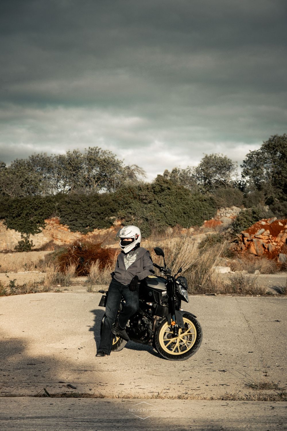 Motorcyclist in white helmet and gray jacket sitting on a black and gold motorcycle on a rural road under cloudy skies.