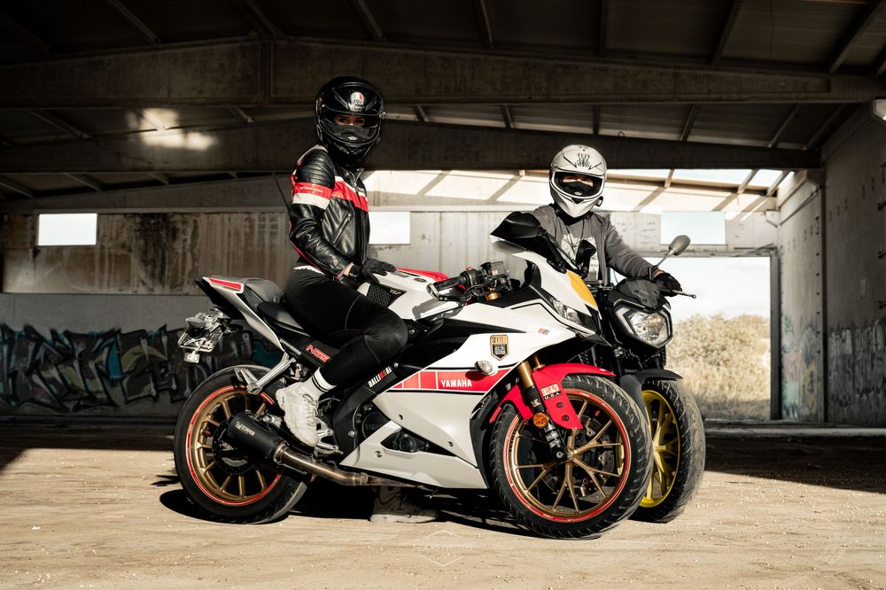Two riders in helmets pose on white and red sport motorcycles inside an industrial building with graffiti walls.