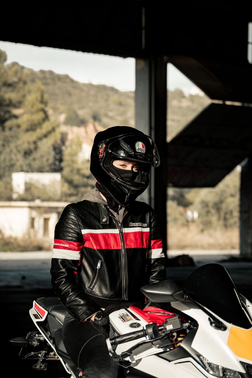 Motorcyclist in black and red leather jacket and full-face helmet sitting on a white sport motorcycle indoors.