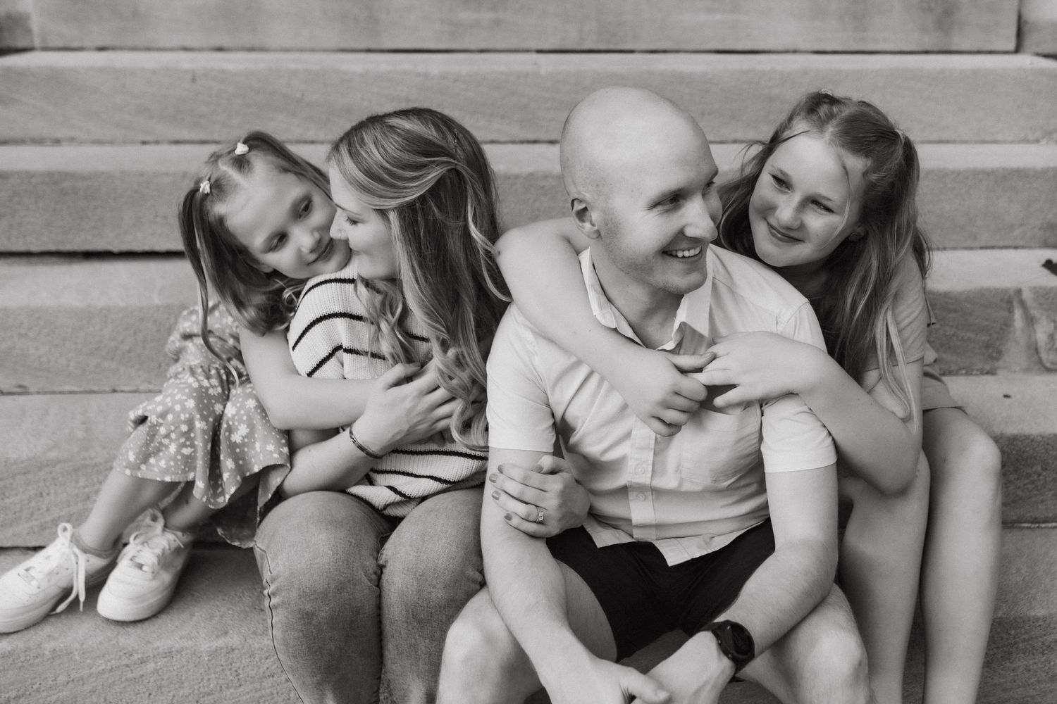 Black and white photo of a bald man sitting on steps with three girls hugging and laughing together in Louisville, KY.