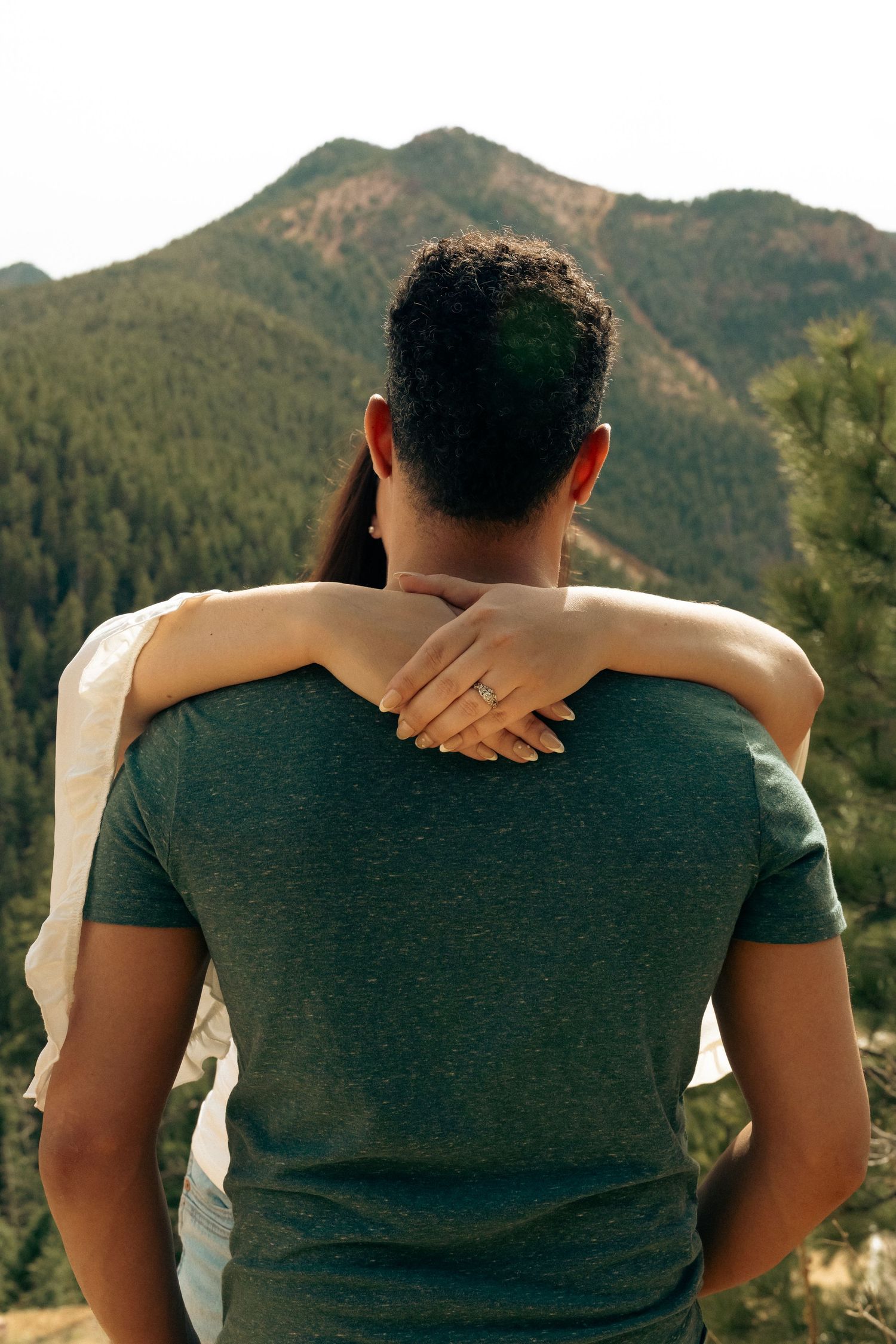 Couple embracing from behind, admiring scenic mountain landscape with lush green trees, woman wearing engagement ring.
