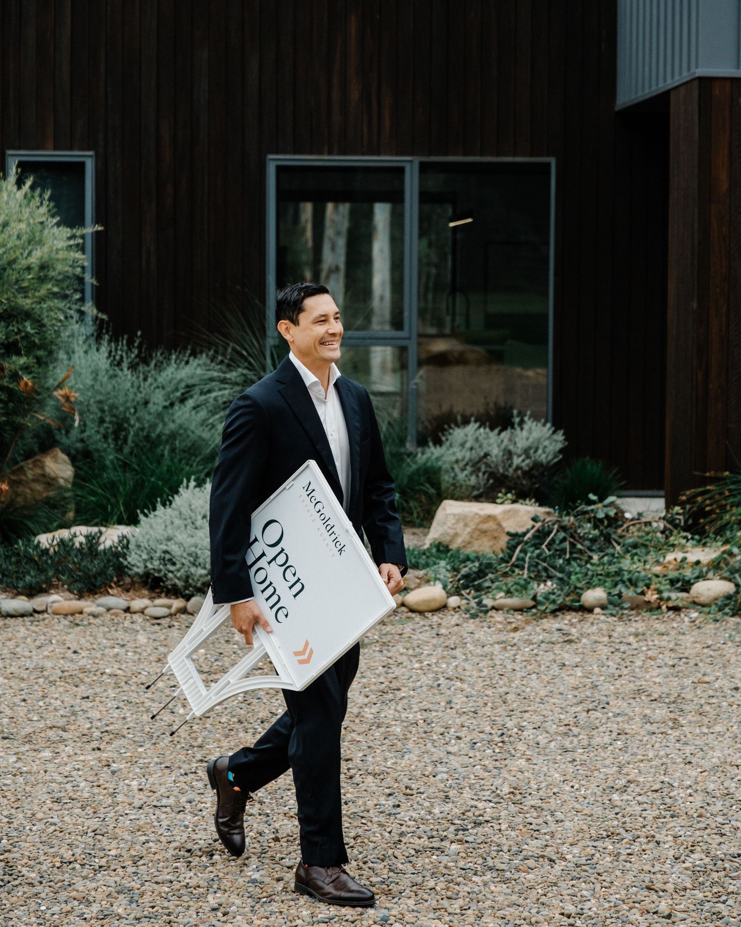 A real estate agent places an Open Home sign on a green lawn in front of a modern Australian home.