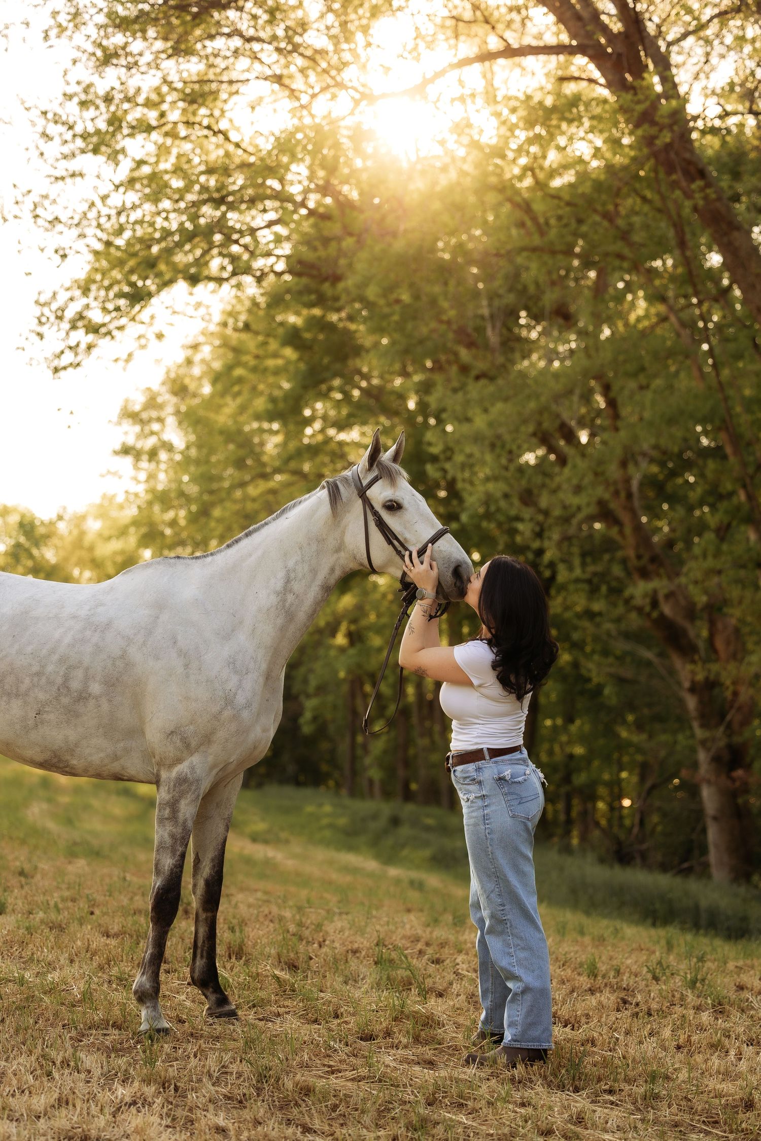 Young woman in white tee and jeans kissing a gray horse at golden hour in a lush green field.