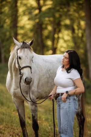 Young woman in white top and jeans stands beside a gray horse in a sunlit forest clearing.