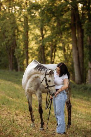 Woman in white tee and jeans tenderly bonding with a dapple gray horse in a lush green forest setting.