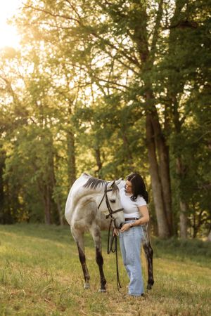 Young woman in white top and jeans tenderly bonding with a gray horse in a sunlit green forest meadow.