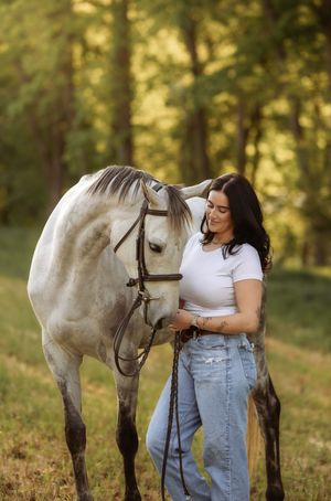 A young woman in a white top and jeans smiles while petting a gray horse in a sunlit green field.