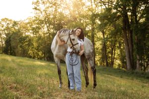 Young woman standing with a gray horse in a sunlit meadow surrounded by lush green trees at golden hour.