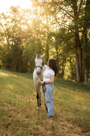 Young woman in white top and jeans stands with a gray horse in a sunlit green field at golden hour.