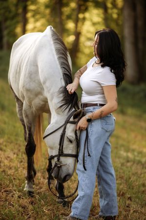Woman in white top and jeans stands beside a white horse in a sunlit forest setting.