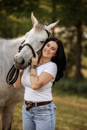 A smiling woman with dark hair in a white t-shirt and jeans lovingly embraces a white horse outdoors.